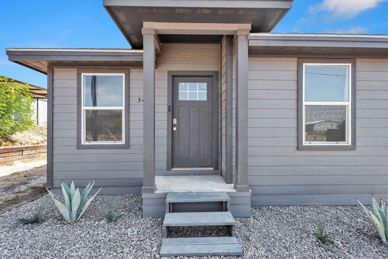 Gray modern home exterior with covered entry, steps, gravel landscaping, and agave plants under a blue sky.