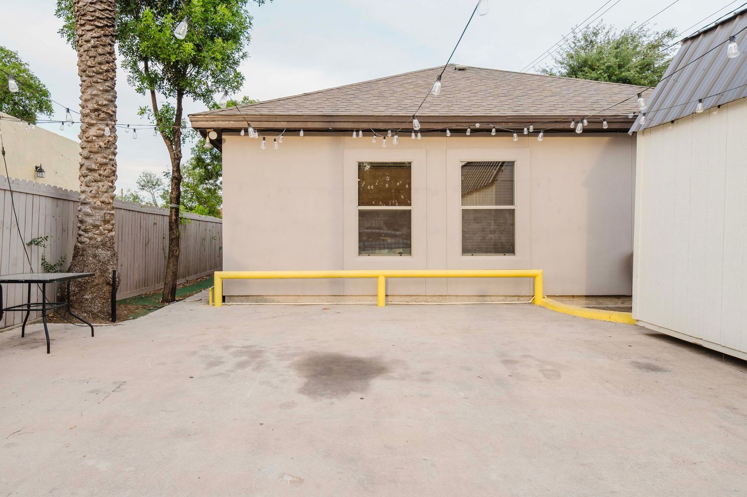 A small beige stucco building with two windows and yellow safety bollards in front of a concrete parking area.