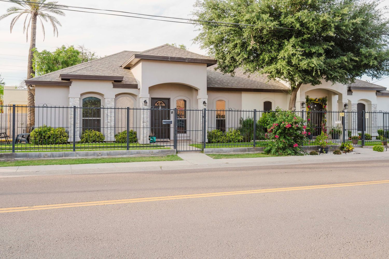 Single-story beige stucco house with wrought iron fence and palm tree along paved street in residential neighborhood.