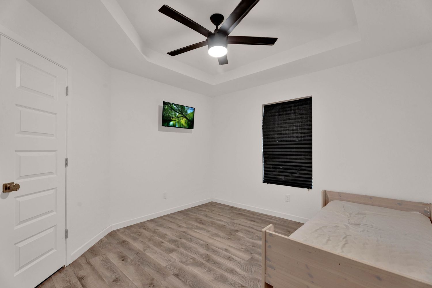 Minimalist bedroom with light wood floors, tray ceiling, black ceiling fan, wall-mounted TV, and a window with dark blinds.