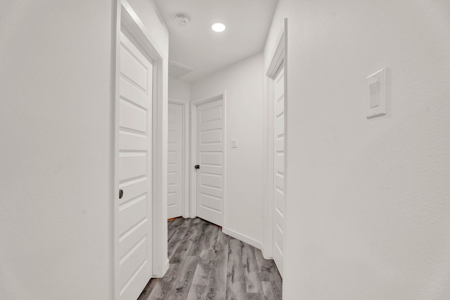 Bright white hallway with gray wood-look flooring, multiple white panel doors, and recessed lighting in a modern home.