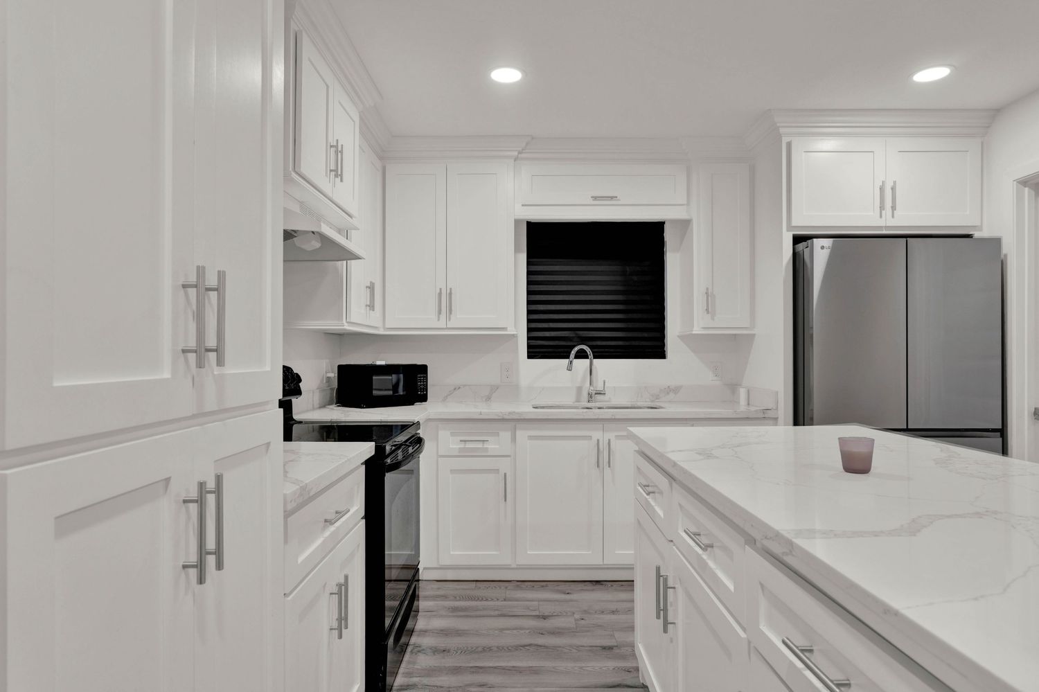 Modern white kitchen with quartz island, stainless steel fridge, black appliances, and shaker cabinets on hardwood floors.
