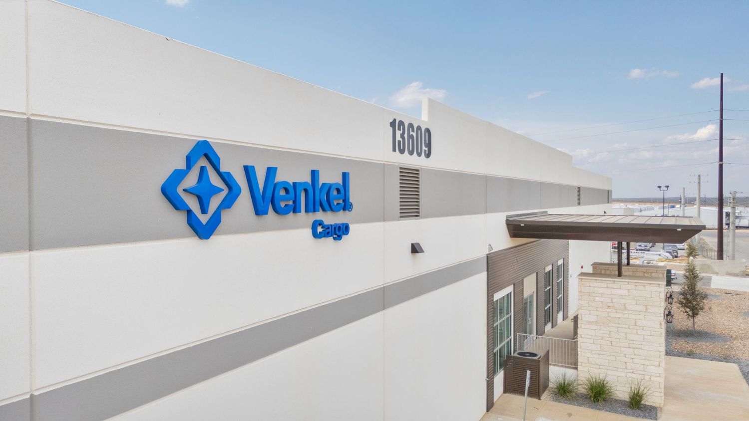 Large industrial warehouse building with white exterior walls and blue signage visible under clear blue sky.