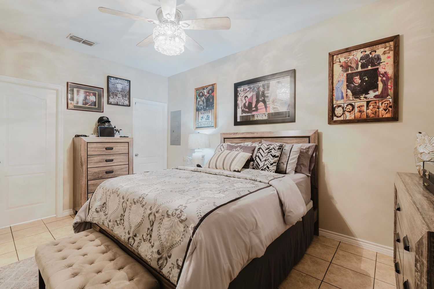 Cozy bedroom with beige bedding, tiled floors, framed wall art, and a wooden dresser under a ceiling fan.
