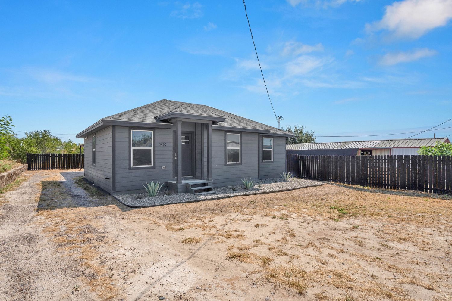 Gray bungalow home with fenced backyard, gravel driveway, and desert landscaping under a blue sky.