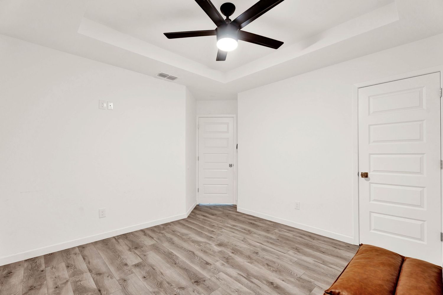 Bright empty bedroom with tray ceiling, black ceiling fan, and light hardwood floors.