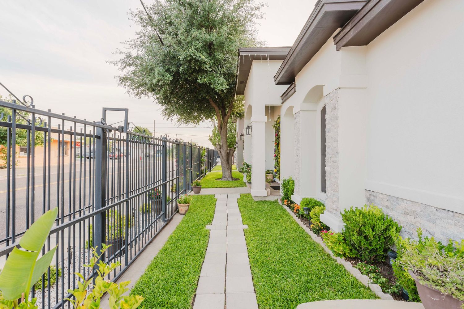 Narrow walkway with green lawn and metal fence beside white stucco building with landscaped shrubs and tree.