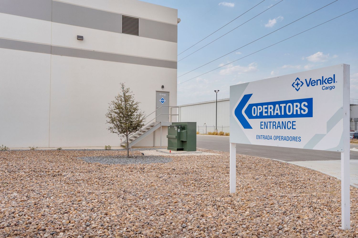 A blue and white Operators Entrance sign stands in front of an industrial building with gravel landscaping.