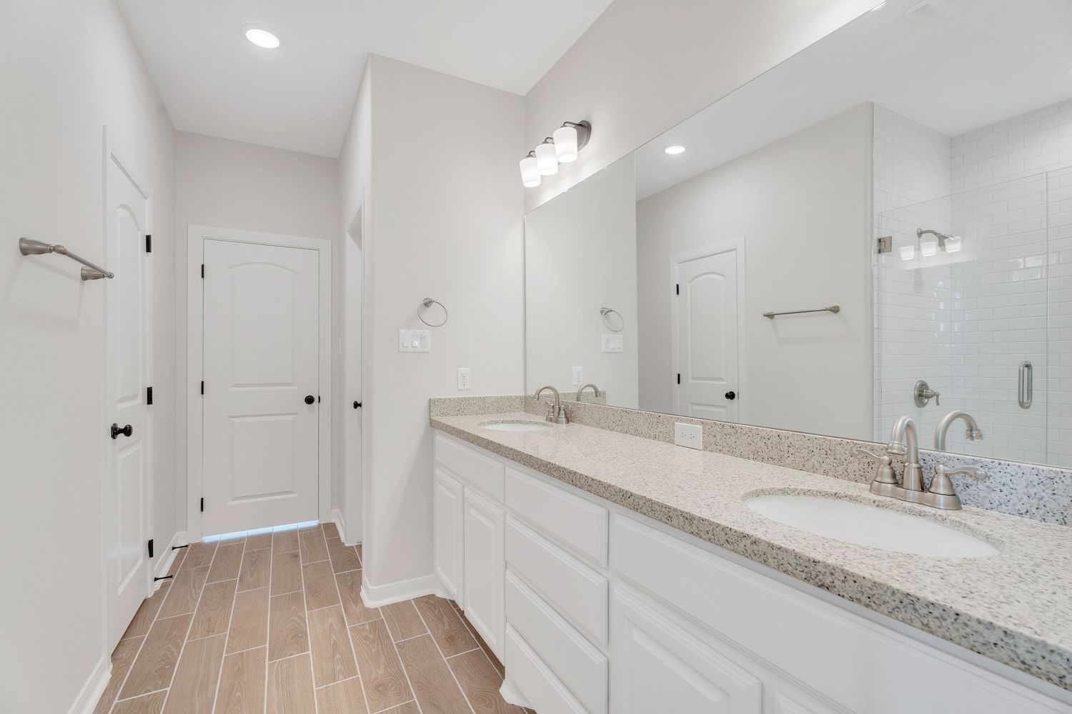 Modern white bathroom interior with granite countertops, recessed lighting, and wood-look tile flooring.