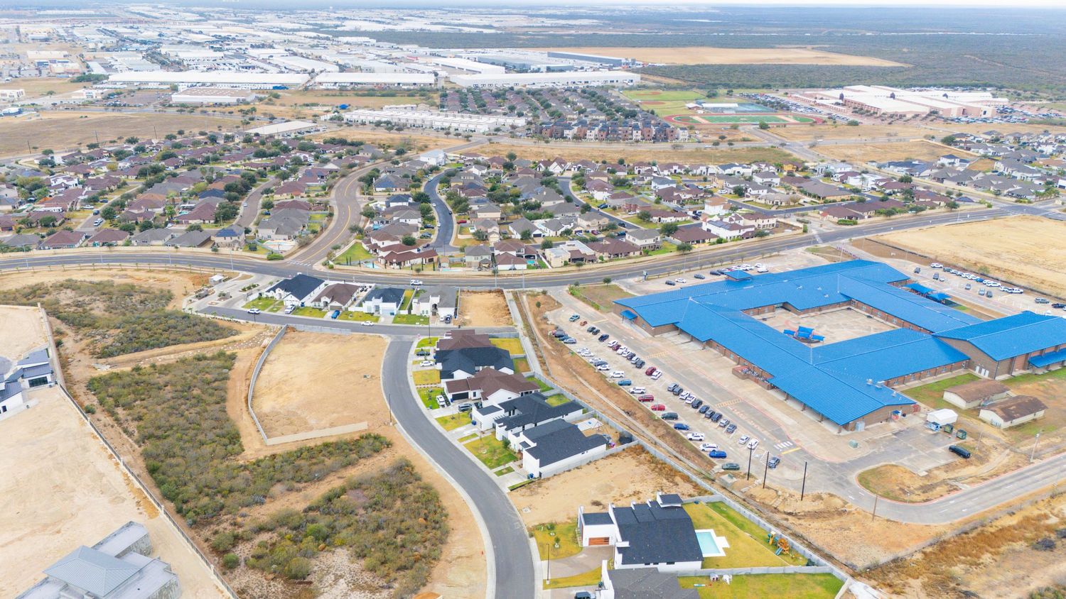 Aerial view of a suburban development with curved roads and industrial buildings near open fields.