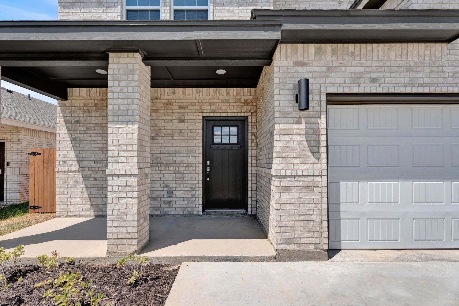 Modern home exterior with light brick, black front door, covered porch, and attached garage on concrete driveway.