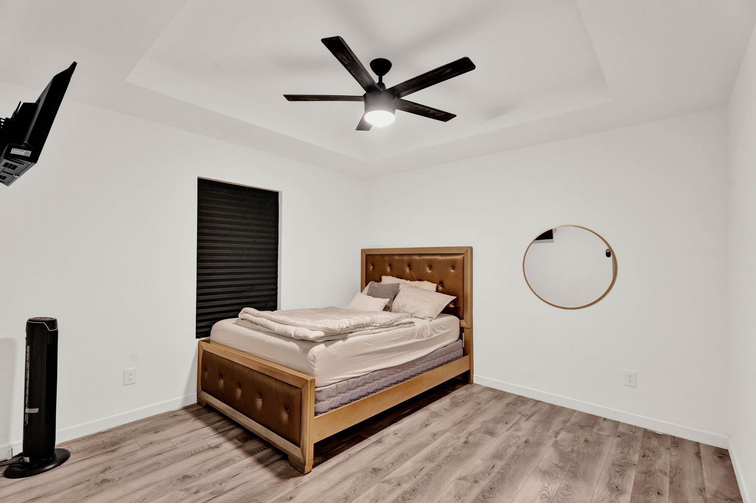 Modern bedroom with wooden bed frame, ceiling fan, hardwood floors, and minimalist white walls with a round mirror.