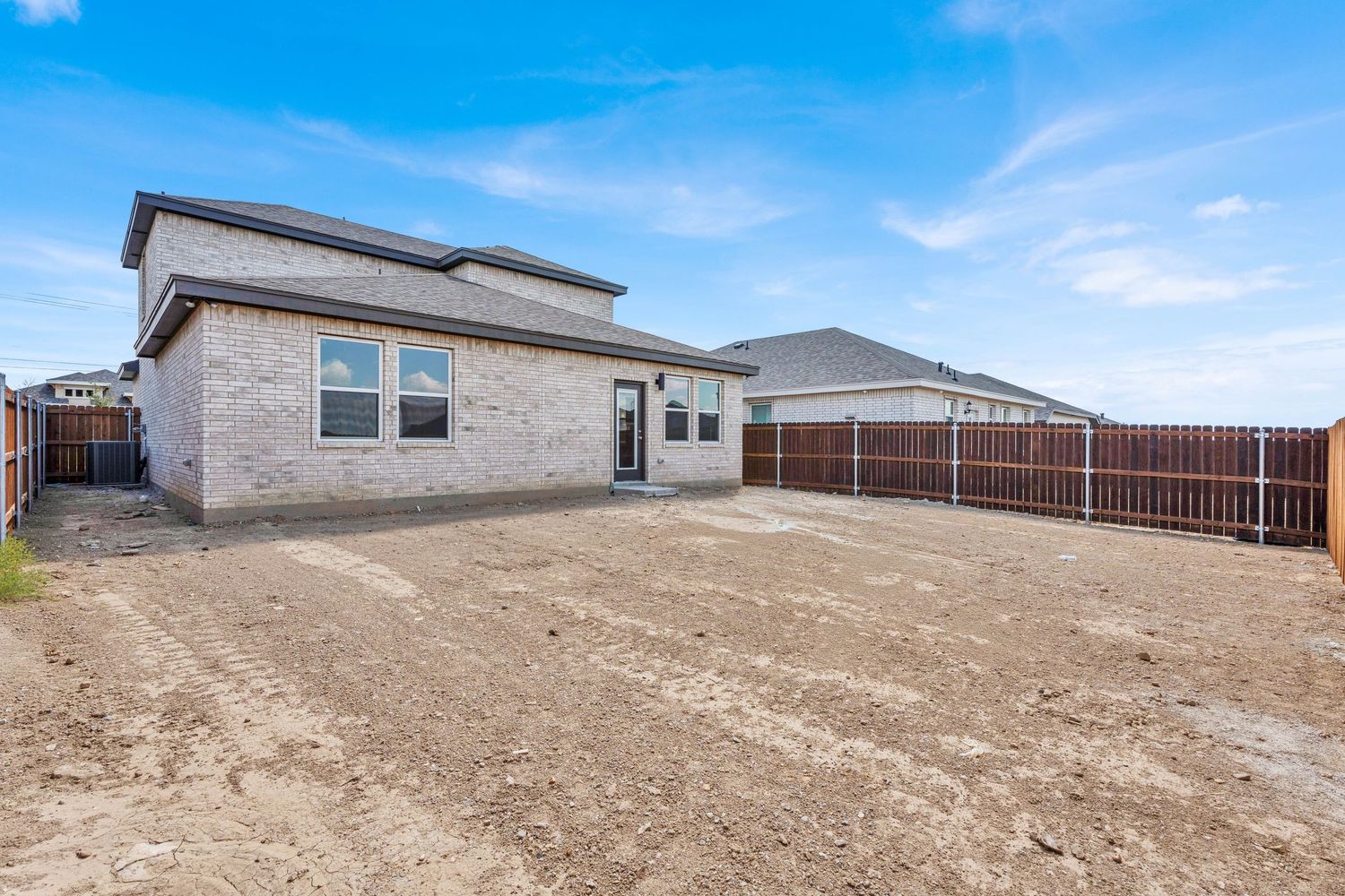 New brick home with expansive dirt backyard, wood privacy fence, and clear blue sky in a suburban neighborhood.