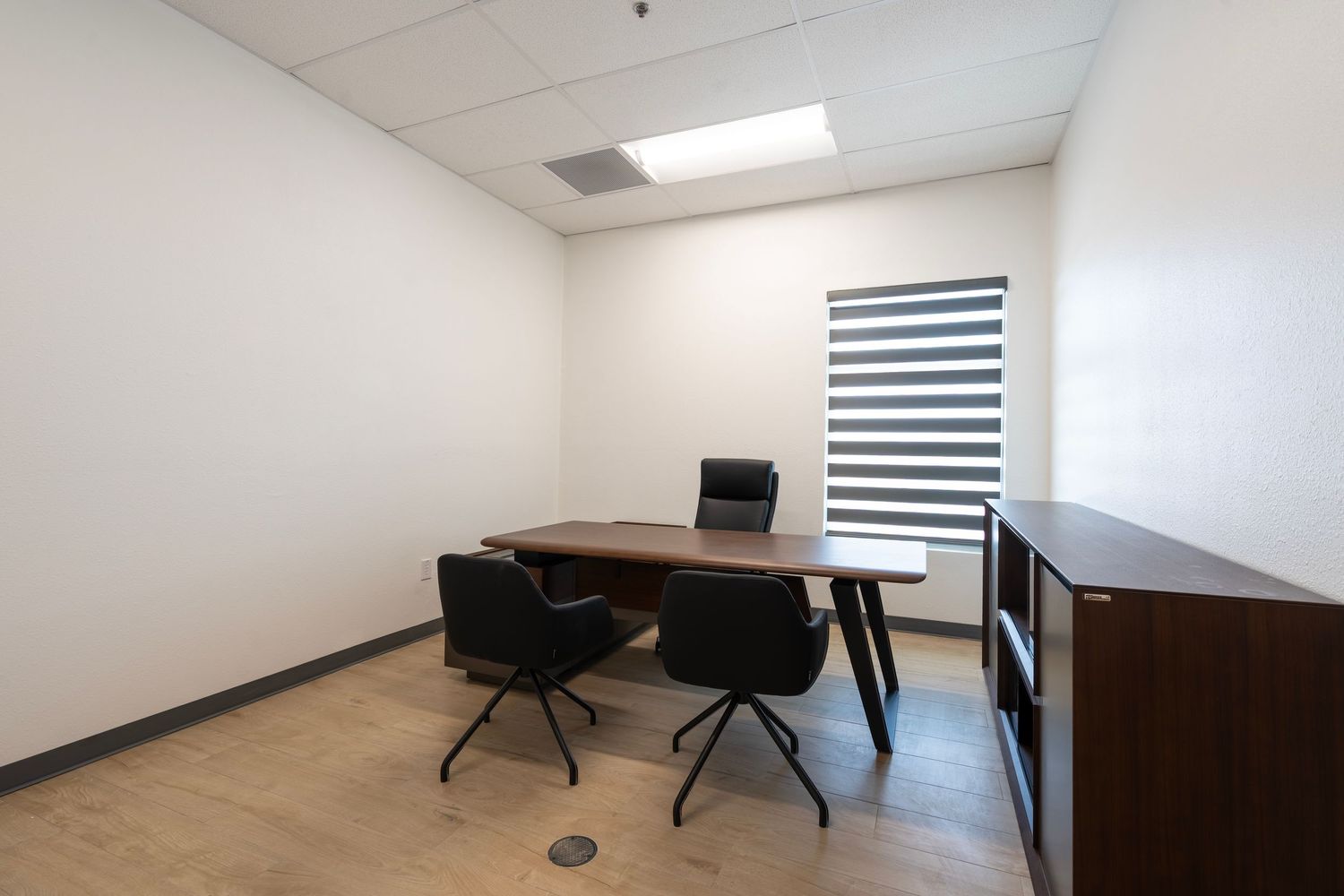 Modern minimalist office with a wooden desk, black chairs, white walls and zebra-style window blinds.