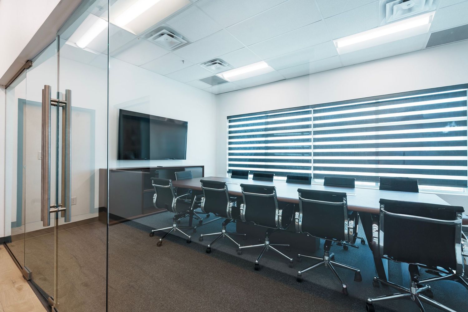 Modern conference room with glass walls, black striped window blinds, and sleek office chairs around a long meeting table.