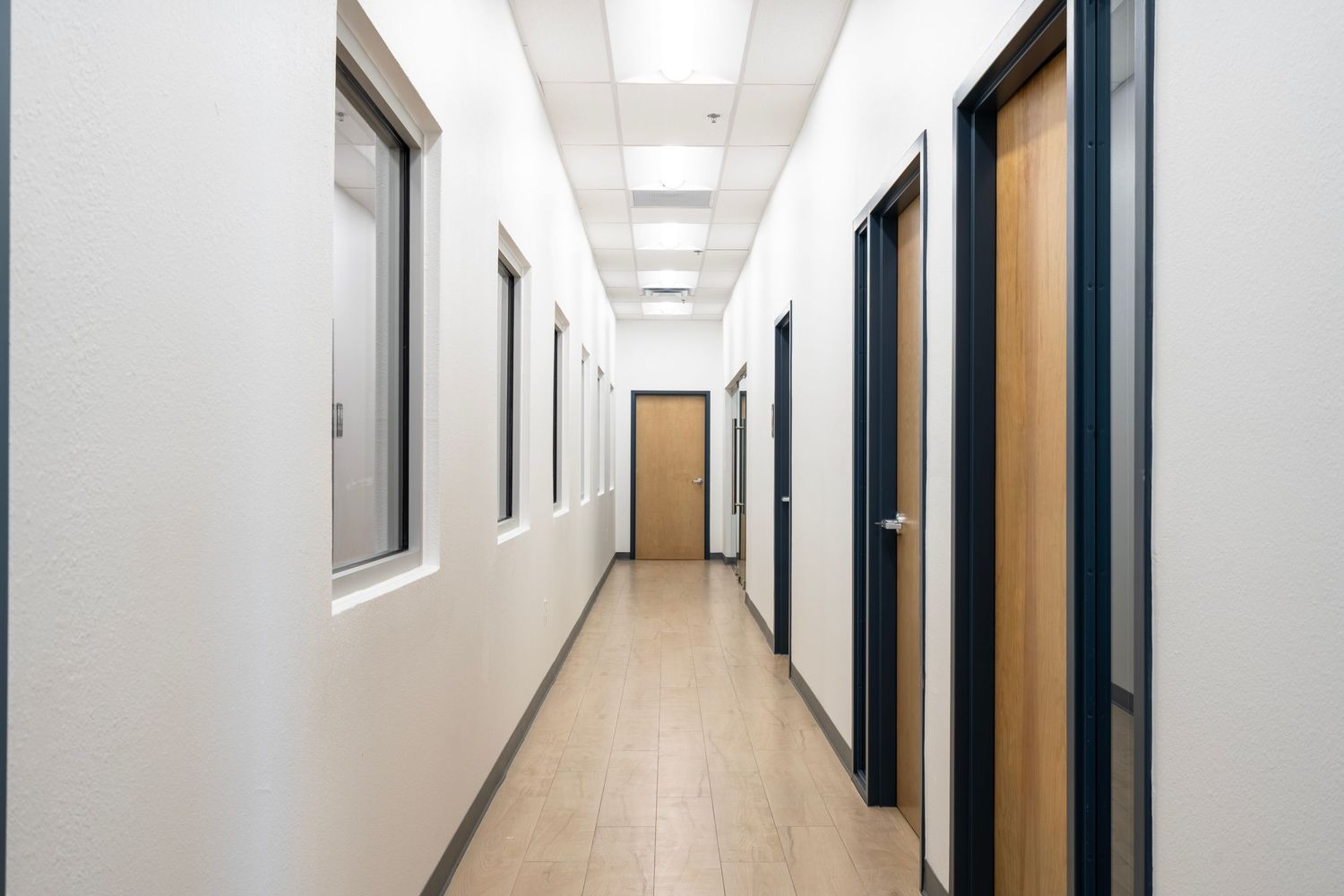 Long office hallway with fluorescent lighting, dark doors on both sides, and beige tiled flooring leading to yellow door.