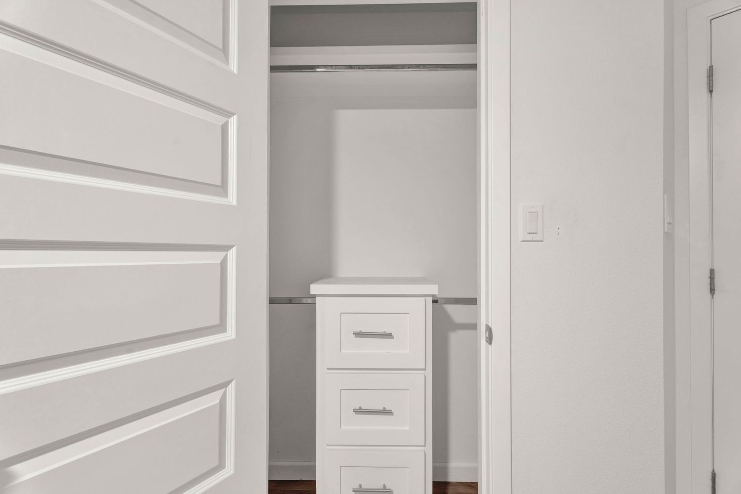 White walk-in closet with built-in shelving, hanging rod, and small white dresser unit.
