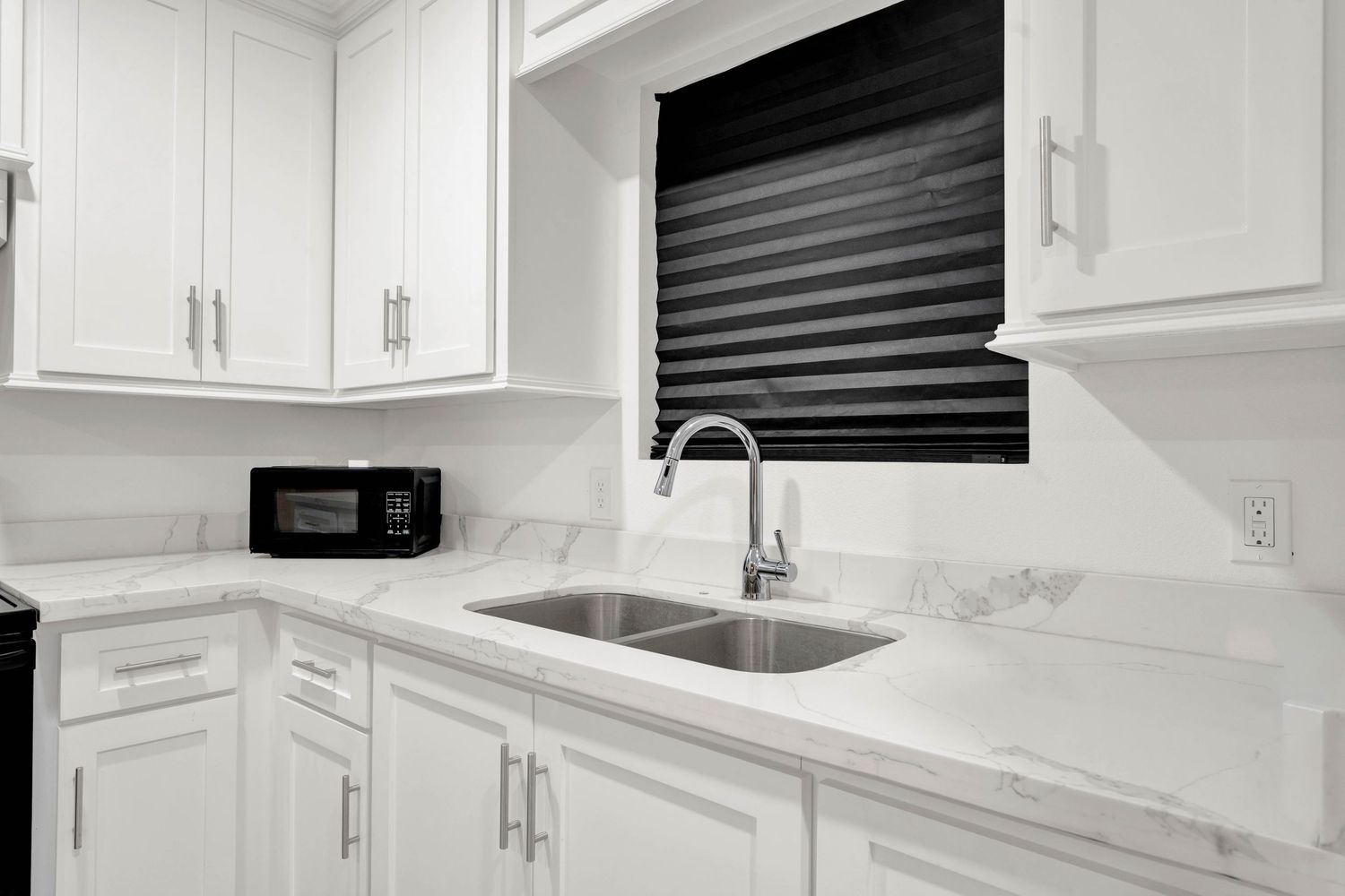 Modern white kitchen with marble countertops, stainless steel sink, white cabinets, and black microwave near window.
