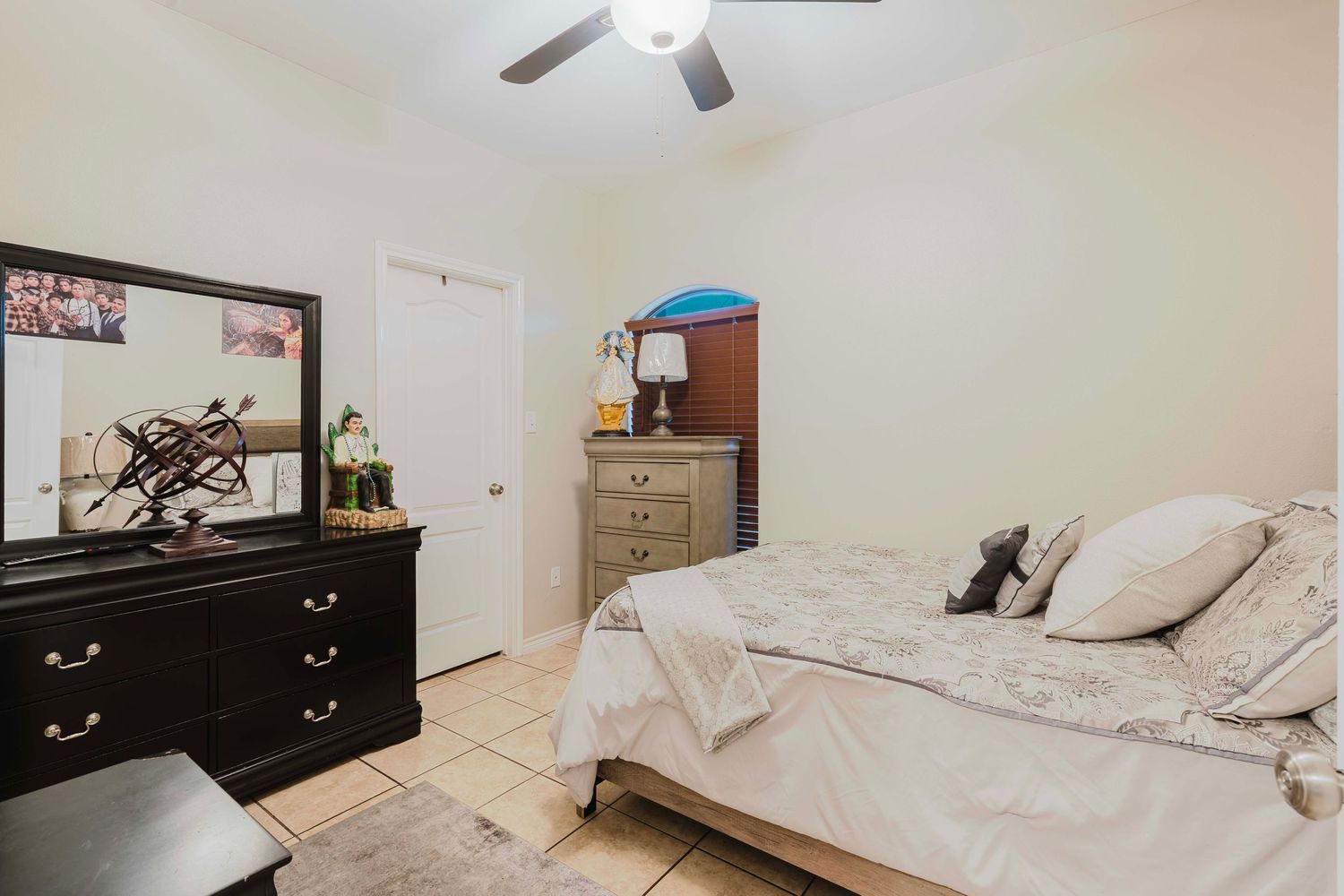 A bedroom with white bedding, dark dresser, TV mounted on wall, and ceiling fan in view.