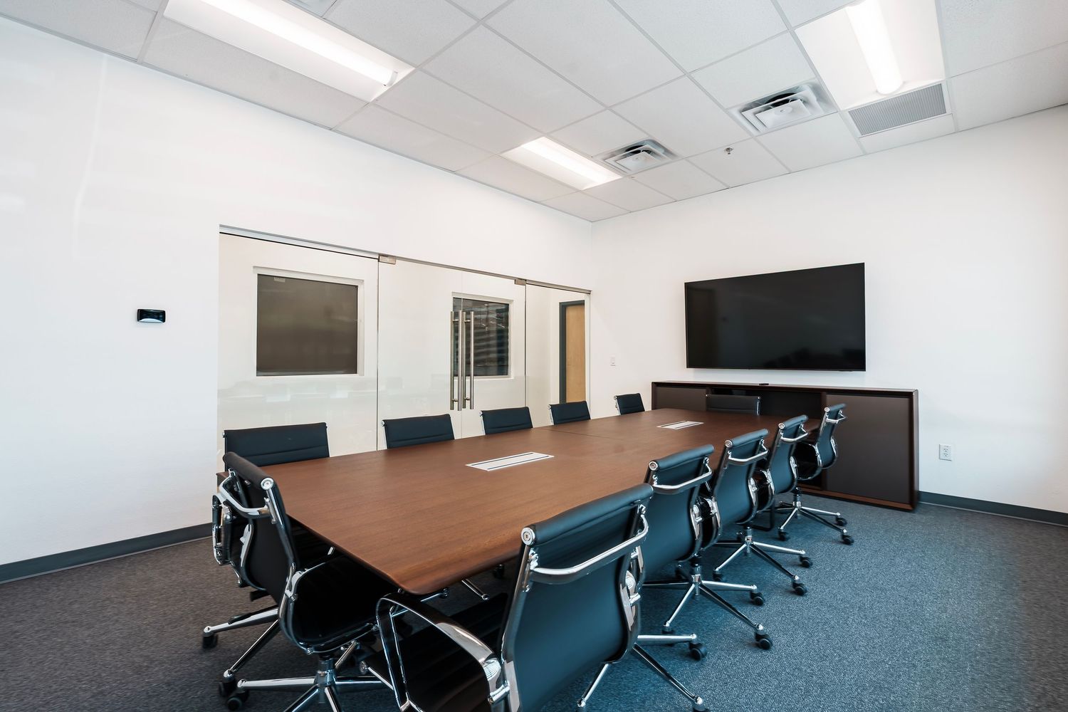 Modern conference room with wooden table, blue office chairs, mounted TV screen and white walls.