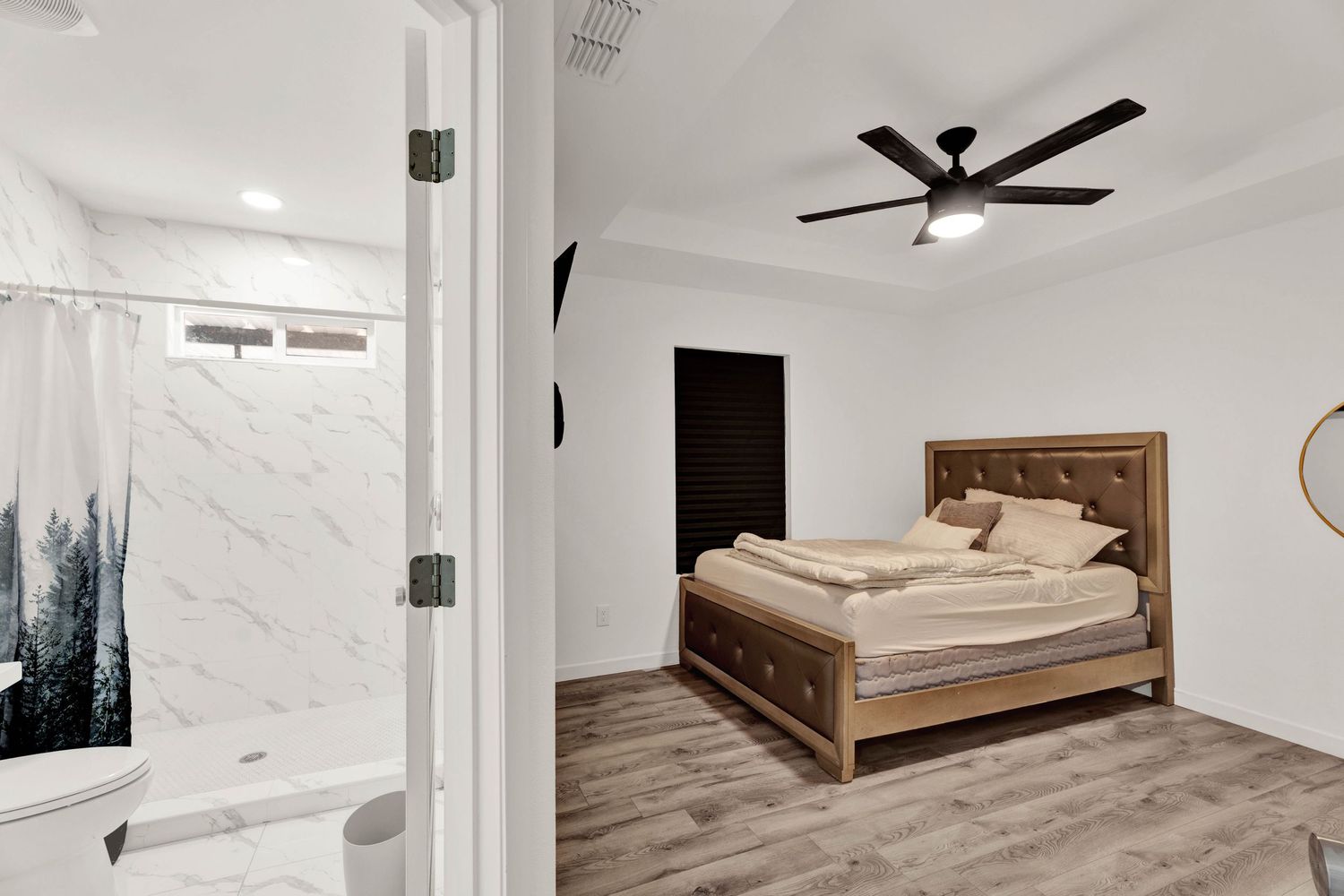 Spacious bedroom with ceiling fan, hardwood floors, upholstered bed, and marble shower visible.