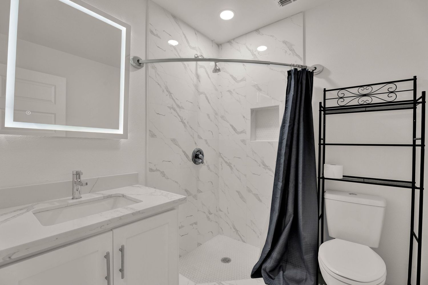 Modern white bathroom with marble shower, black curtain, vanity with LED mirror, and over-toilet storage shelf.