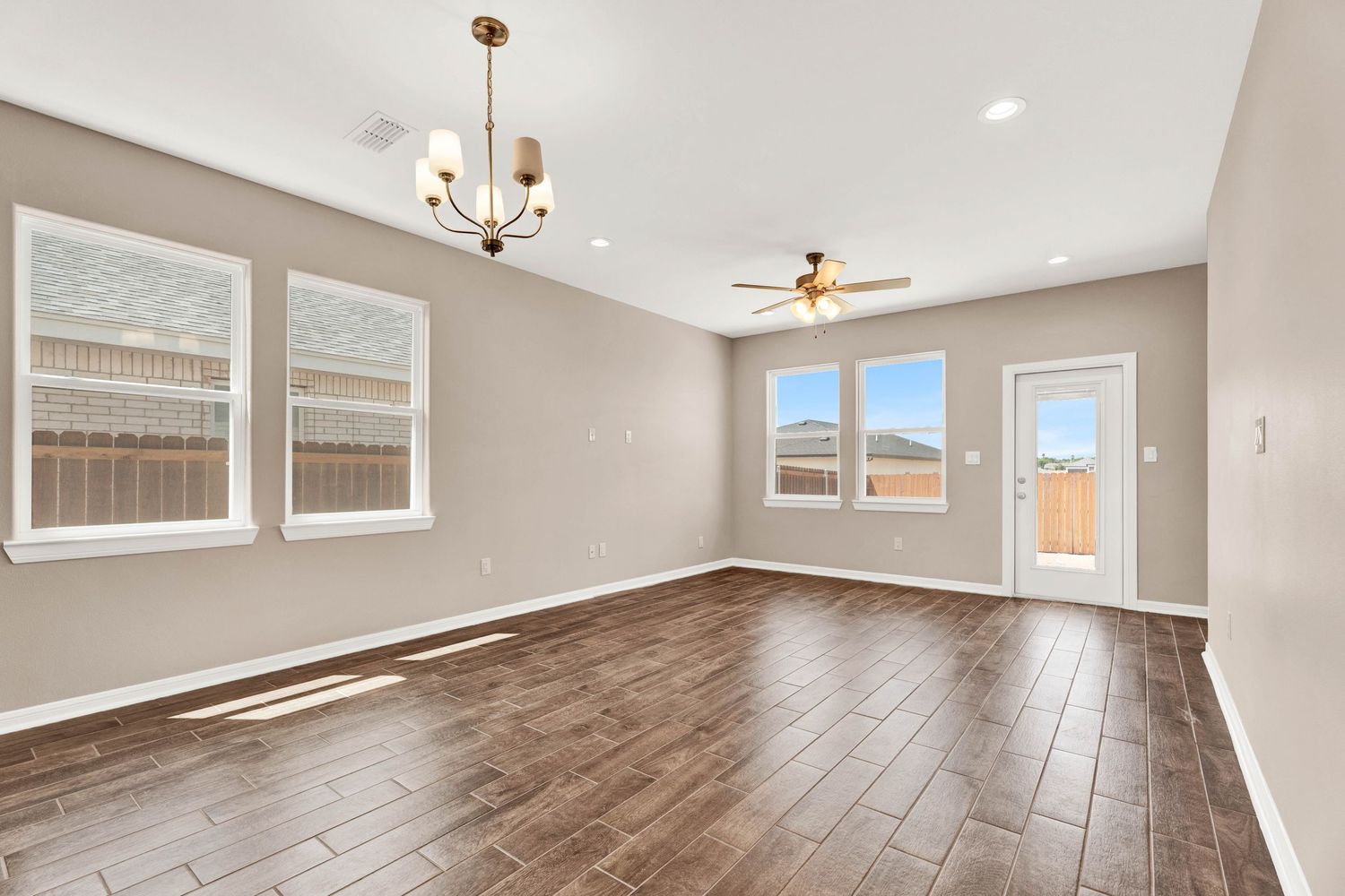 Bright open living area with wood-look floors, neutral walls, ceiling fan, chandelier, and large windows letting in natural light.