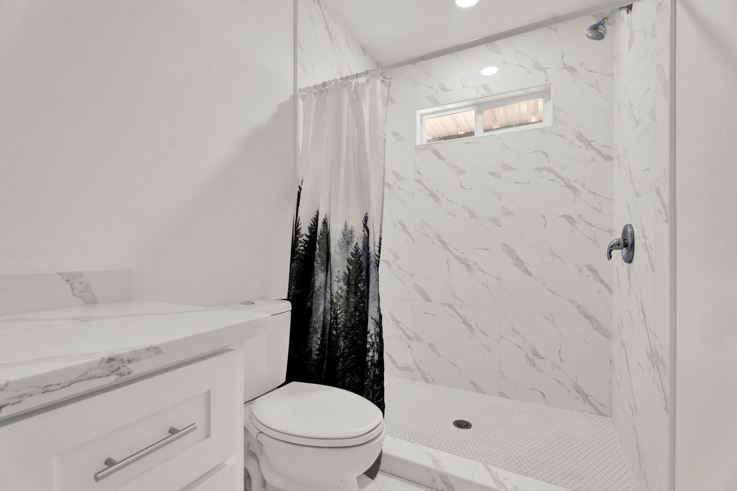 Clean white bathroom with marble shower surround, forest print curtain, toilet, and vanity.
