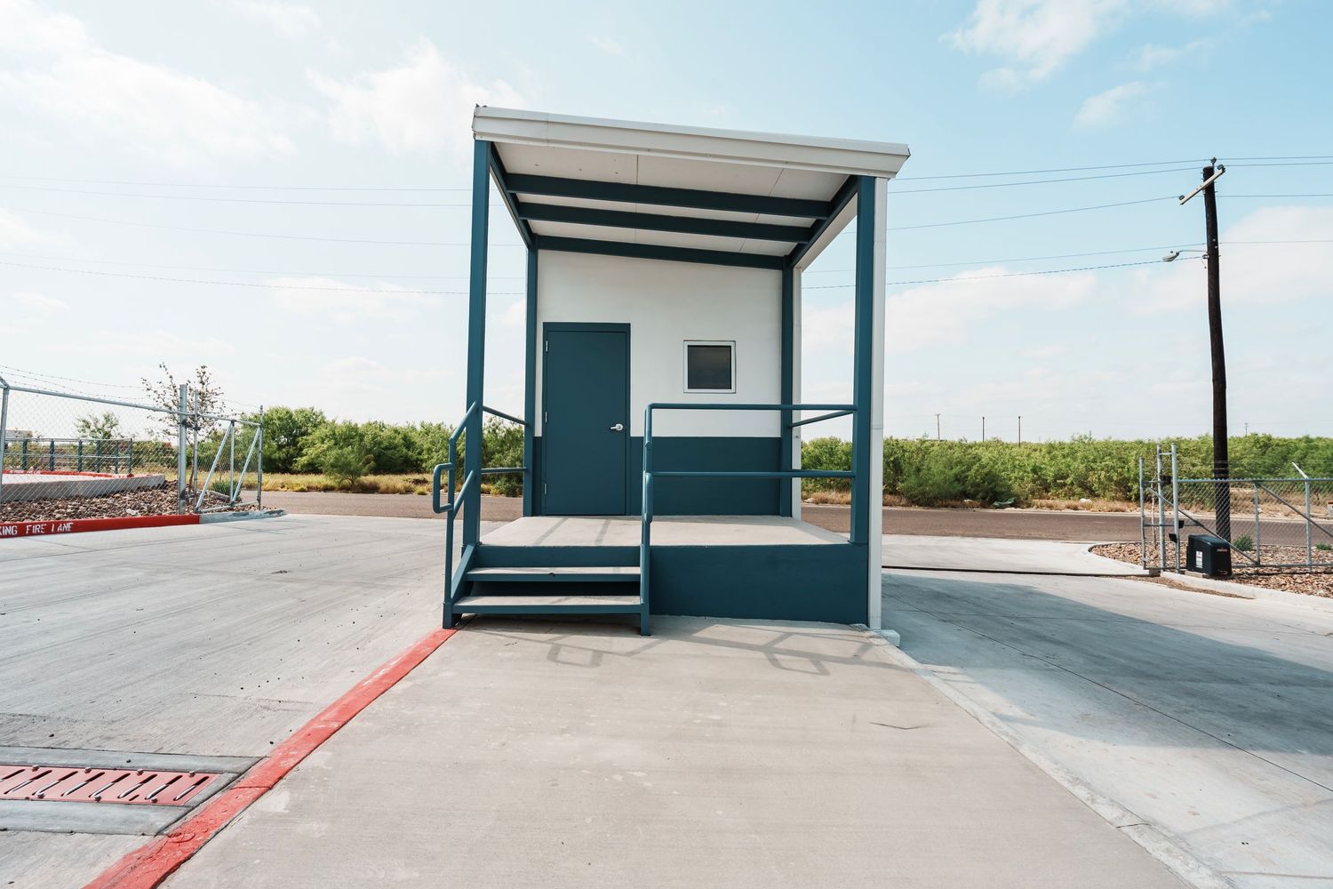 A small modern guard booth or security kiosk with teal accents stands at an entrance to a paved area under blue skies.