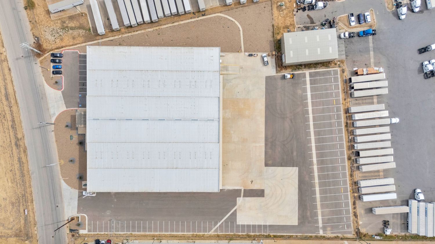 Aerial view of a commercial warehouse facility with white roof and adjacent parking area for semi-trucks and trailers.