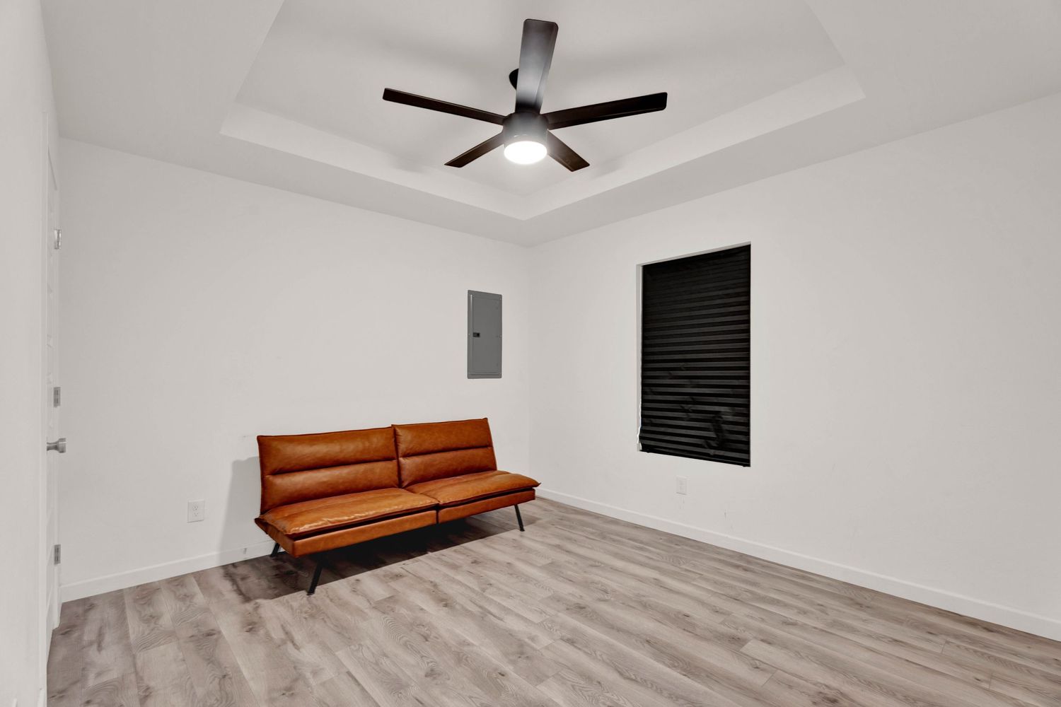 Modern bedroom with brown leather bench, ceiling fan, tray ceiling, and hardwood floors in a bright white room.