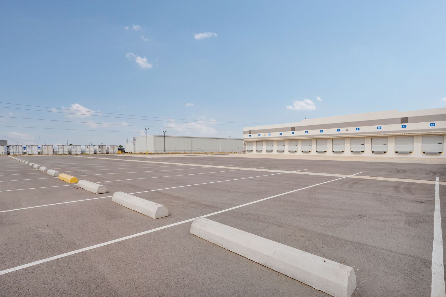 Empty parking lot with white line markings and concrete barriers under blue sky with scattered clouds.