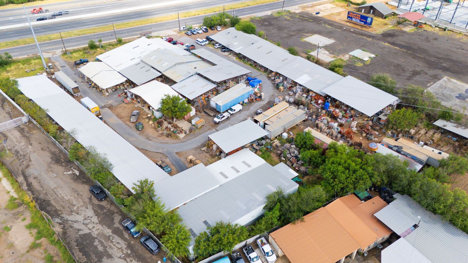 Aerial view of industrial warehouse complex with metal roofs and storage yard surrounded by paved roads and dirt lots.