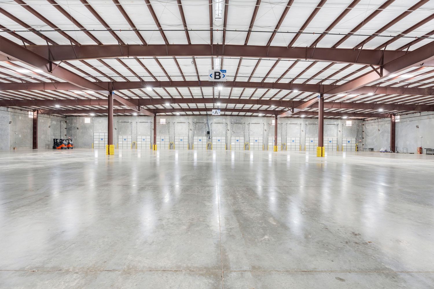 Large empty warehouse interior with metal roof beams and concrete floor showing sequential construction progress.
