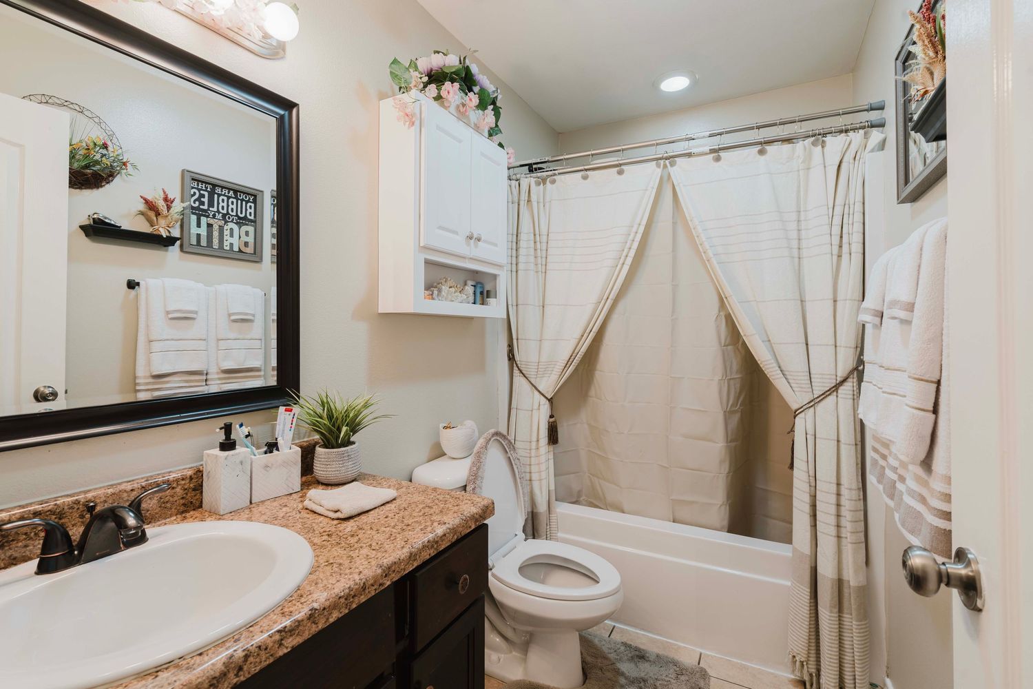 Modern bathroom featuring beige shower curtain, granite countertop vanity, and white cabinets with decorative plants.