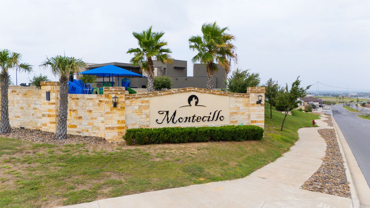 A panoramic view of a residential entrance with palm trees, stone walls, and a winding road.