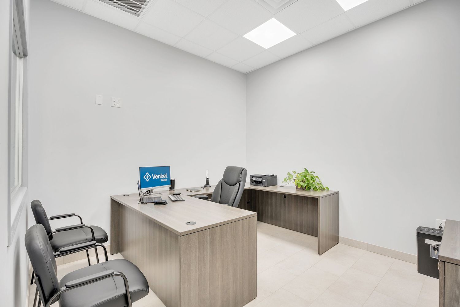 Modern office interior with white walls, wooden desks, black chairs and bright ceiling lighting in a clean workspace.