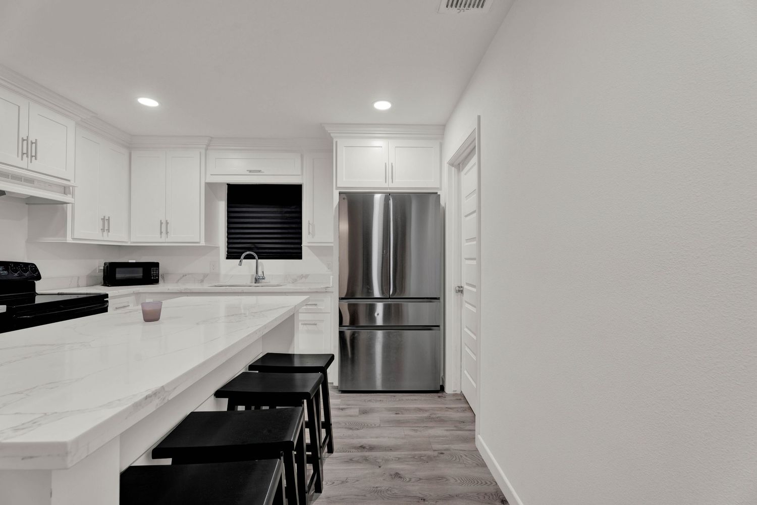 Modern white kitchen with stainless steel refrigerator, quartz countertops, black bar stools, and hardwood floors.