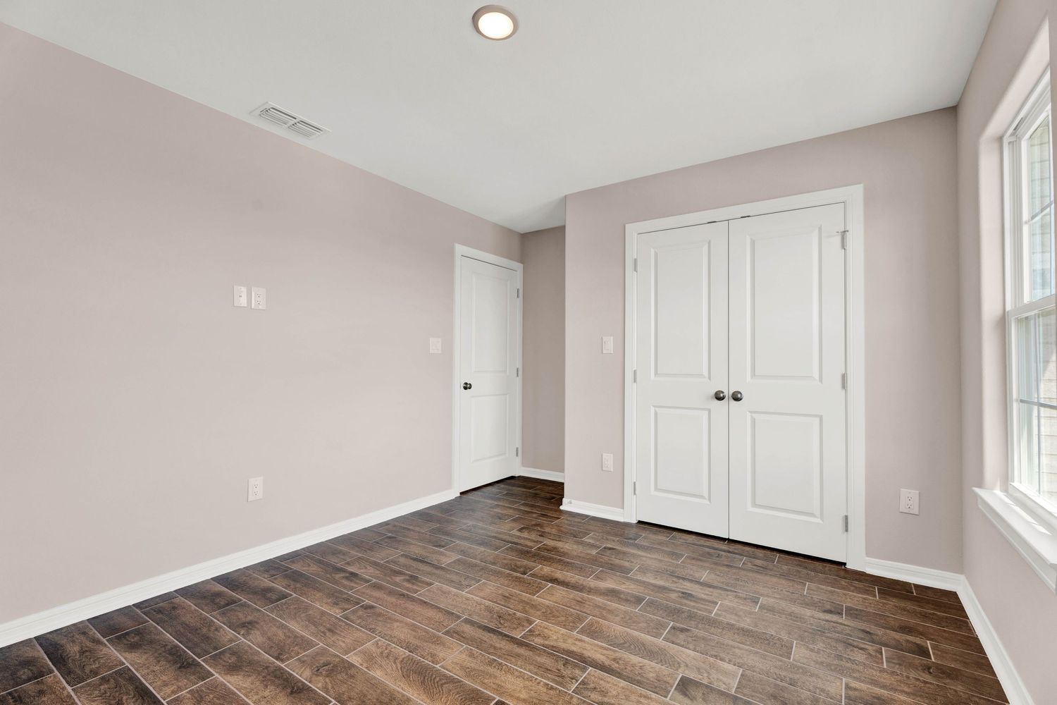 Empty bedroom with dark hardwood floors, pink walls, white double closet doors, and natural light from a window.
