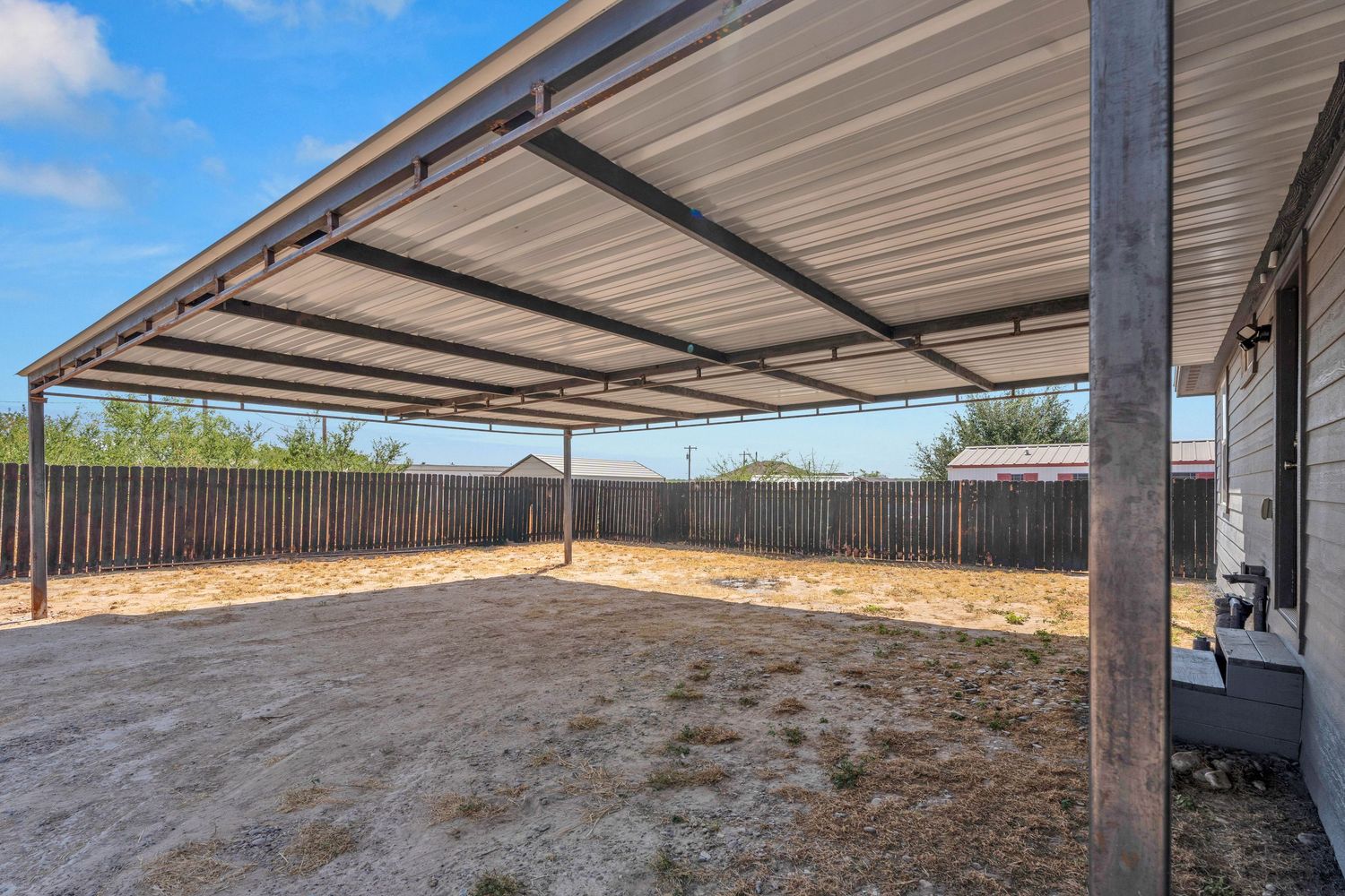 Large covered carport with metal roof attached to home features a spacious dirt yard enclosed by wooden privacy fence.