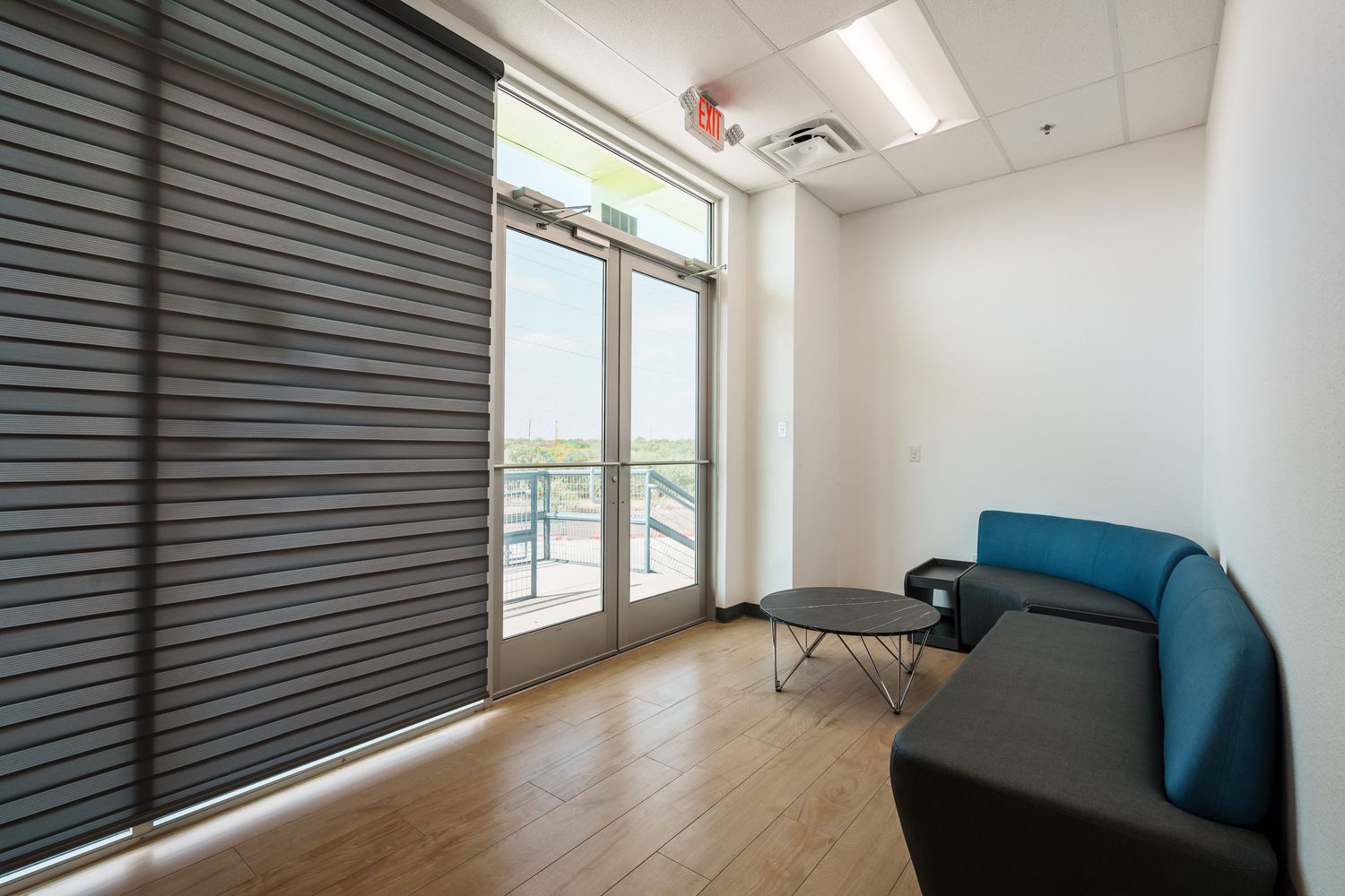Modern office room with floor-to-ceiling window, blue couch, and dark roller blinds features hardwood flooring and ocean view.
