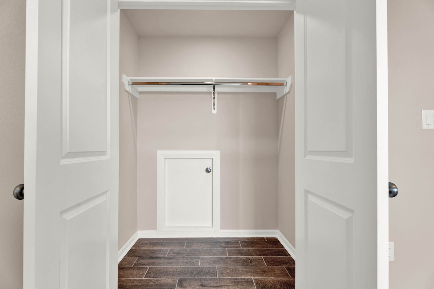 Walk-in closet with white double doors, single hanging rod, and dark wood-look flooring in a neutral beige space.