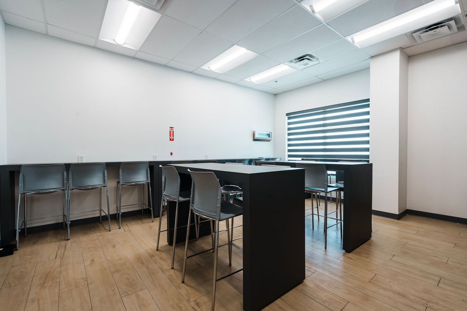 Modern office break room interior with black counter, bar stools, and wooden flooring under fluorescent lighting.