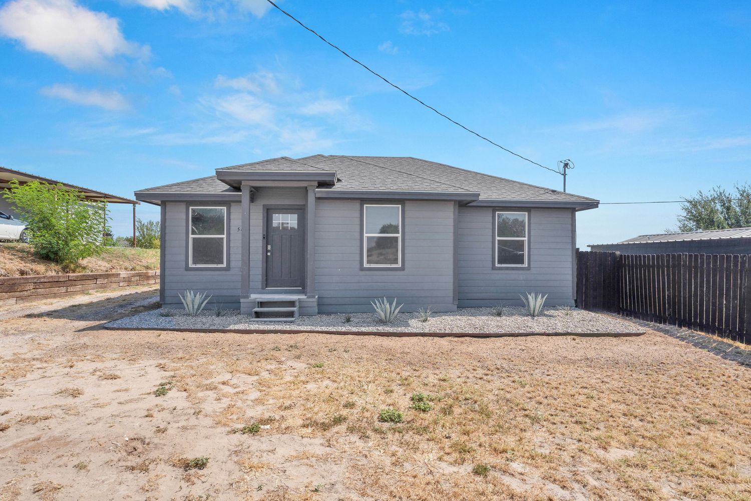 Gray bungalow home with covered entry, large windows, and gravel yard in a sunny residential neighborhood.