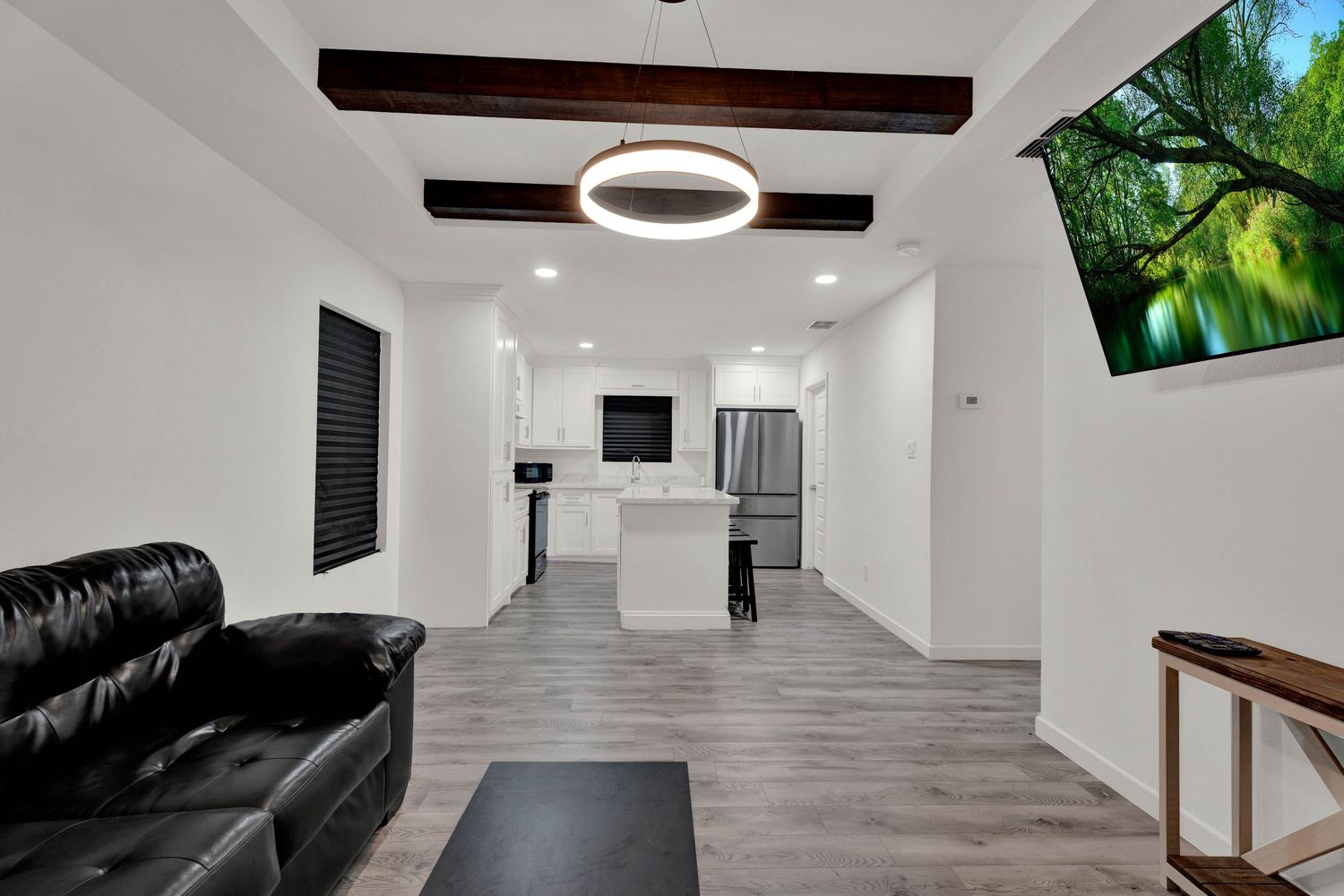 Modern open-plan living room with black leather sofa, wood beam ceiling, circular pendant light, and white kitchen with island.