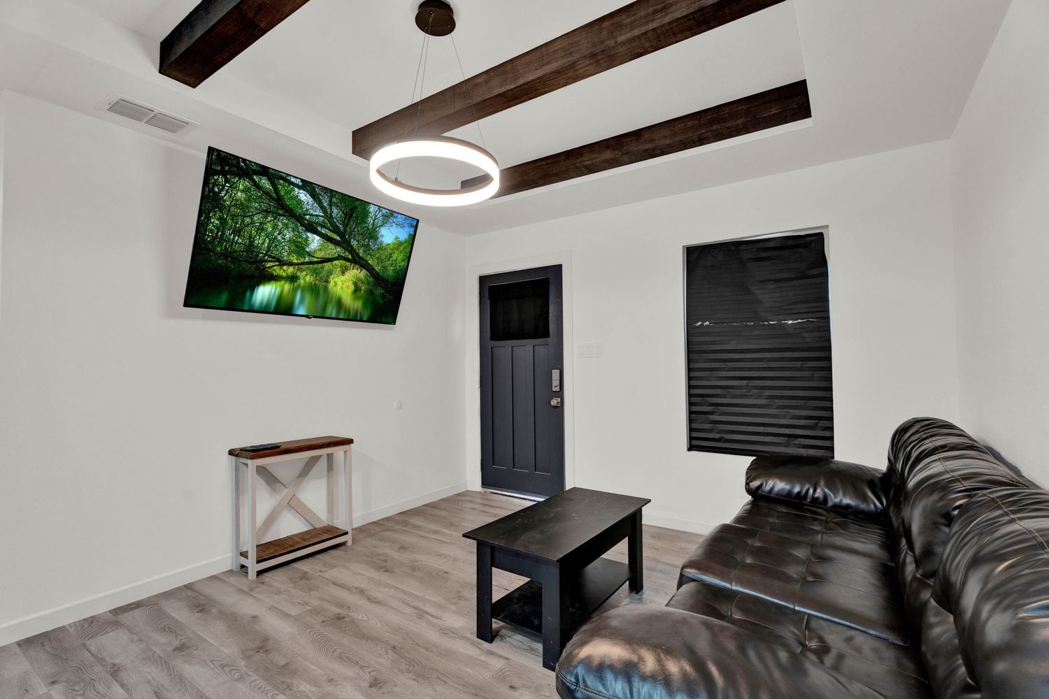 Modern living room with leather sofa, wood beam ceiling, wall-mounted TV, and dark door with black blinds.