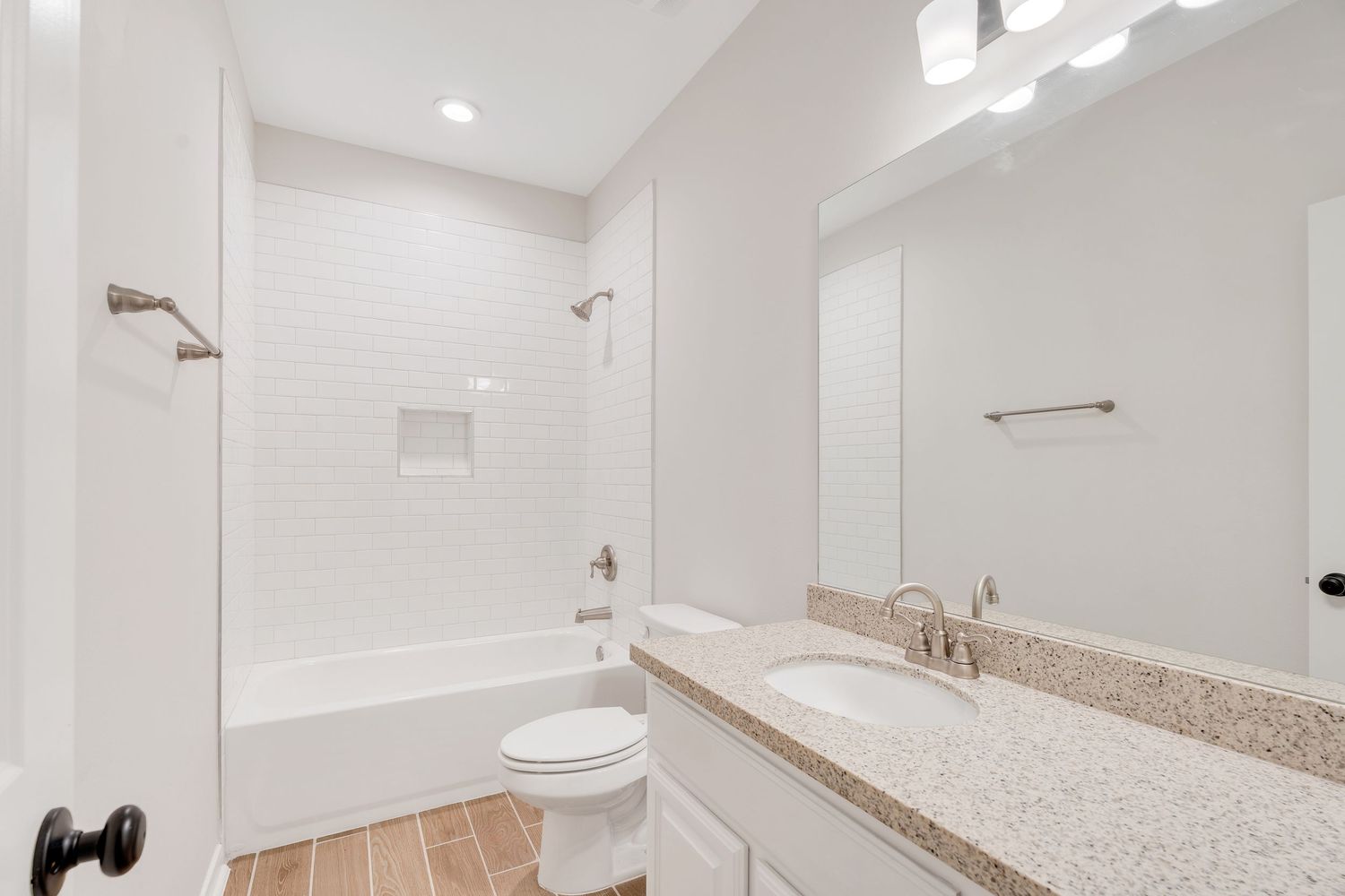 Modern white bathroom with granite countertop, built-in tub, subway tile shower surround and wood-look flooring.