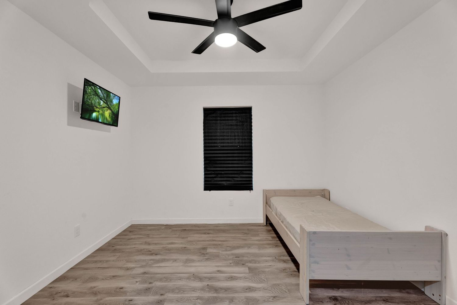 Minimalist bedroom with hardwood floors, ceiling fan, wall-mounted TV, black blinds, and a twin bed frame.