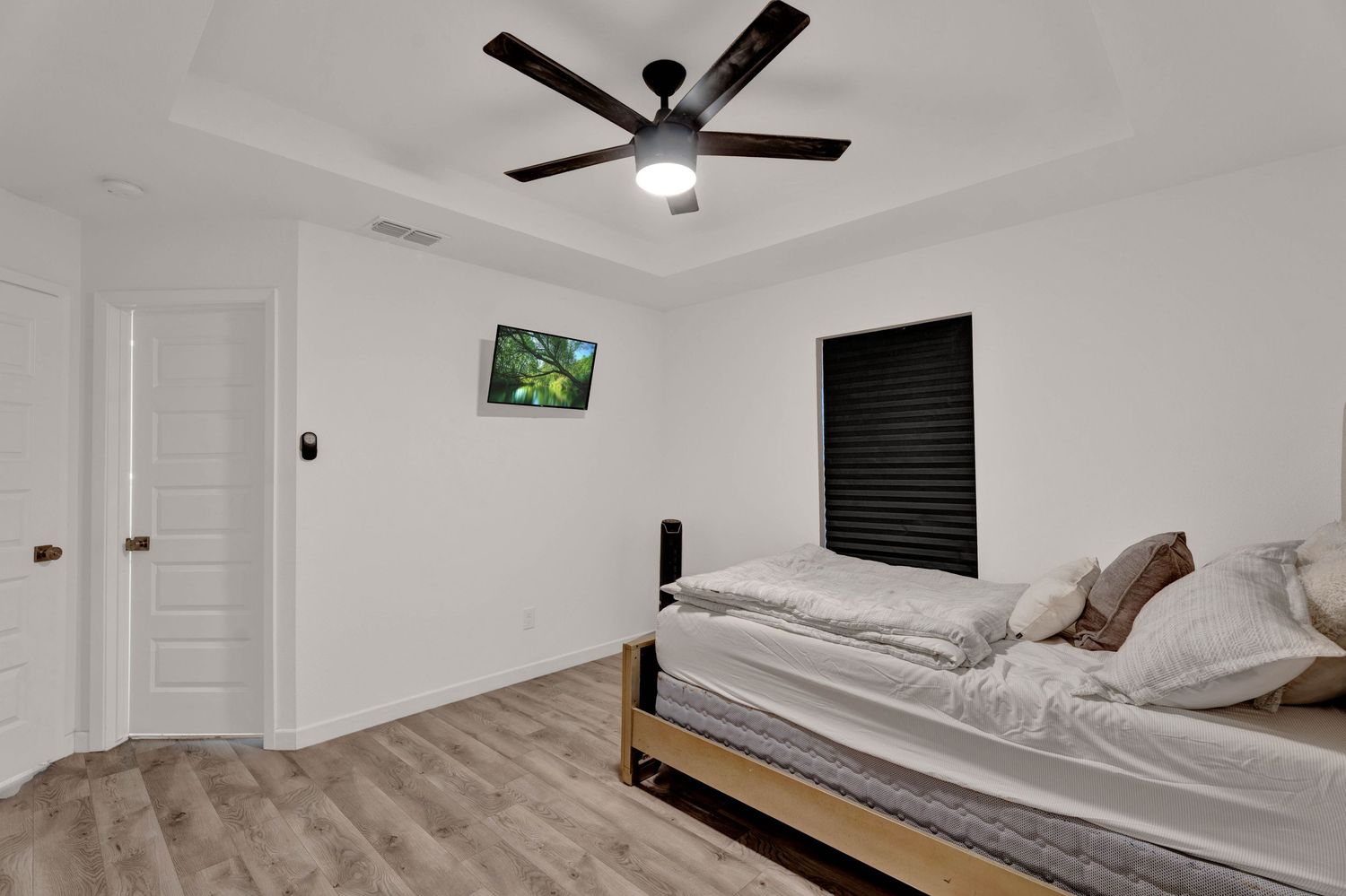 Modern bedroom with ceiling fan, hardwood floors, a bed with neutral bedding, wall-mounted TV, and tray ceiling.