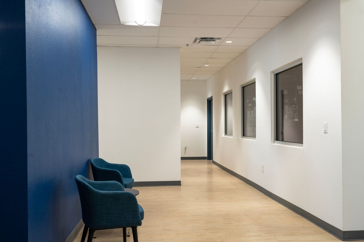 Empty office hallway with blue accent wall and teal chair against white walls and fluorescent lighting.